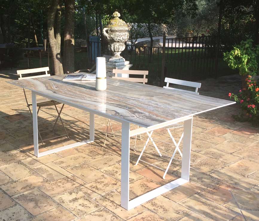 table avec plateau en céramique imitation marbre et pied en métal laqué blanc, sur une terrasse extérieure.
