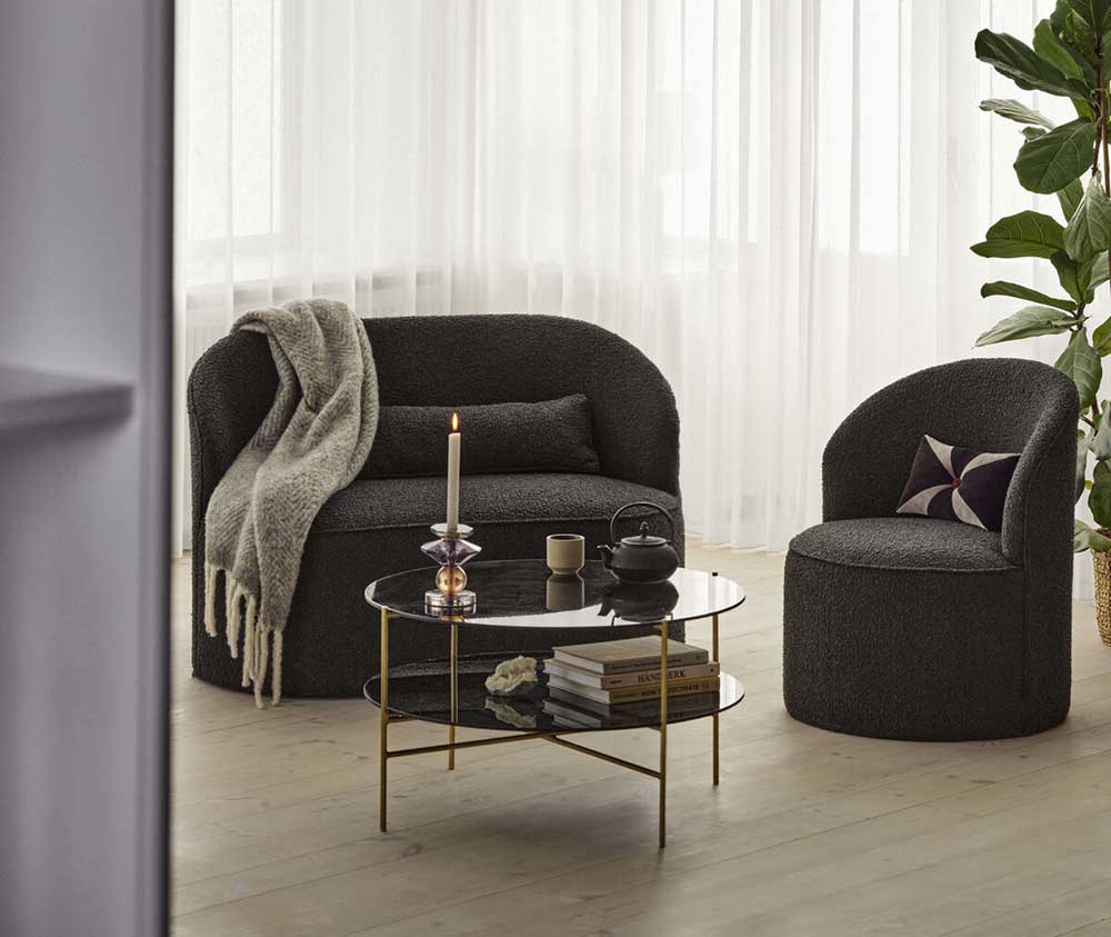 Scène représentant un salon avec parquet et grande fenêtre devant laquelle se trouvent un canapé et fauteuil Lounge en tissu doux bouclé gris charbon