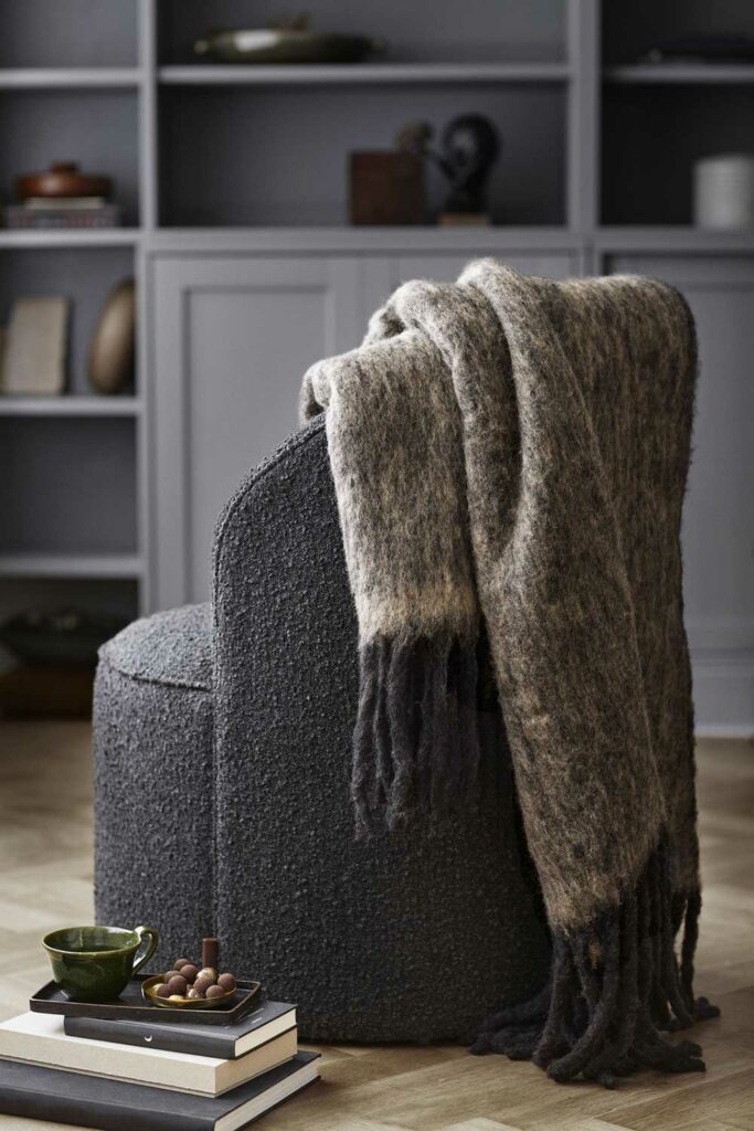 fauteuil lounge en tissu bouclé gris charbon avec formes arrondies. Présentation avec un plaid sur le dossier et des livres au pied du fauteuil. Ambiance Cosy et design.