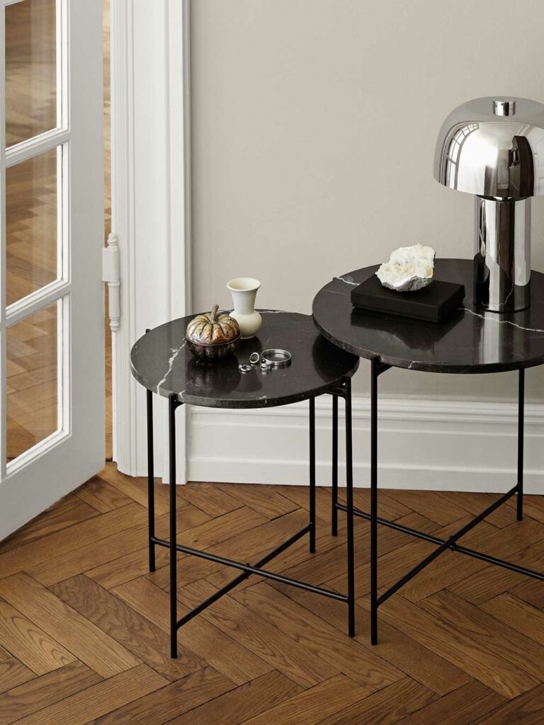 Tables gigognes rondes avec plateau en marbre noir et pied en métal noir. Une table est surmonté d'une lampe chromée. Parquet foncé à chevrons au sol
