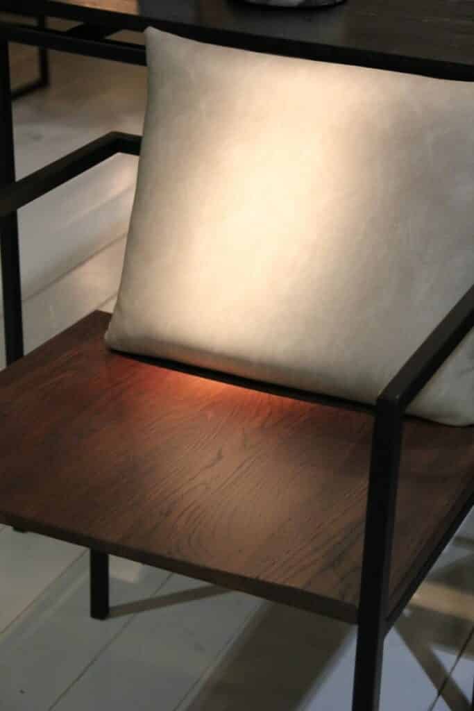 Détail de l'assise en chêne massif, finition Noyer et du coussin en cuir synthétique lisse couleur Army. Le cadre avec accoudoirs est en métal noir.